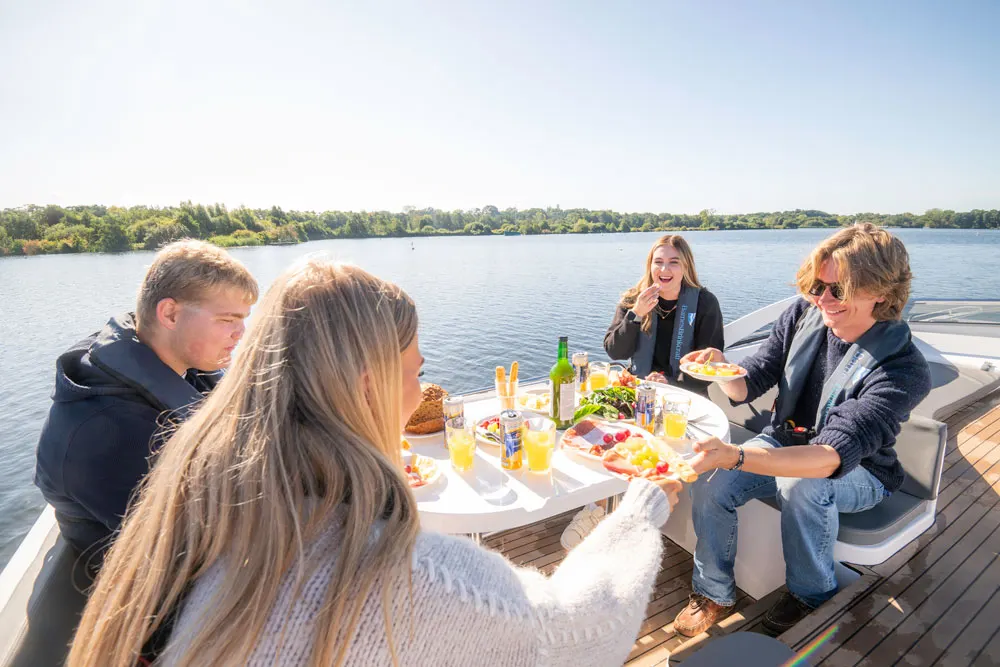 Group of four people dining together at a table on a boat on a calm lake, with wooded shoreline in the background.