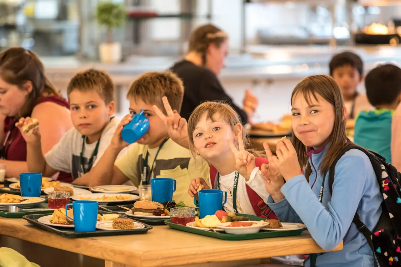 Children eating a meal together at a dining table, some wearing lanyards, with an adult supervisor in the background.
