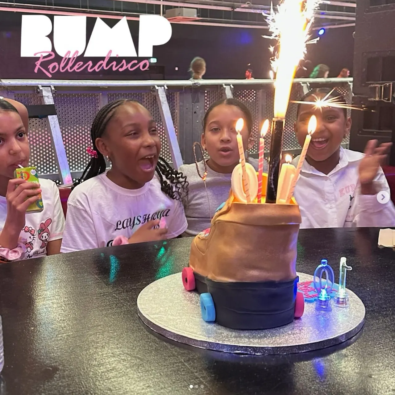 Young children celebrating at a roller disco venue with an illuminated birthday cake on wheels in front of them.