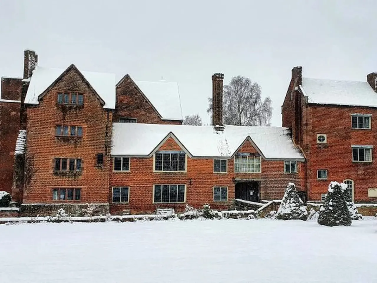 A large red brick Tudor mansion with snow-covered roofs and grounds, set against a winter landscape with bare trees.