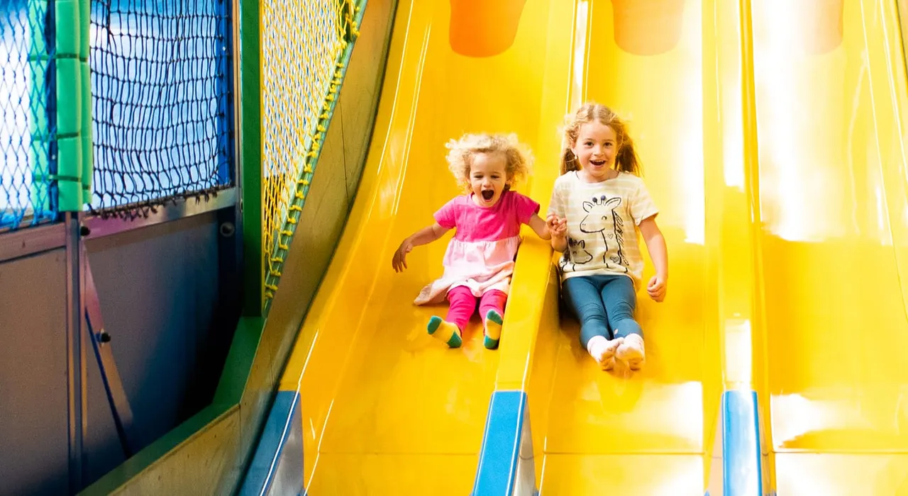 Two young children sliding down a bright yellow indoor slide, smiling with excitement.