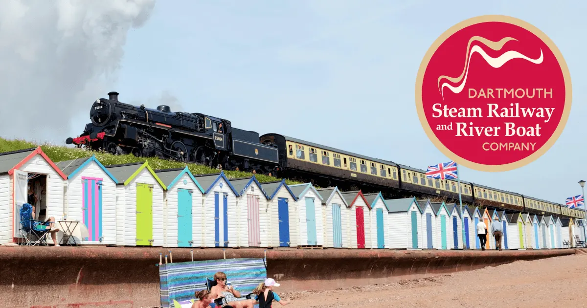 Steam train travelling above colourful beach huts on sand, with visitors relaxing below.