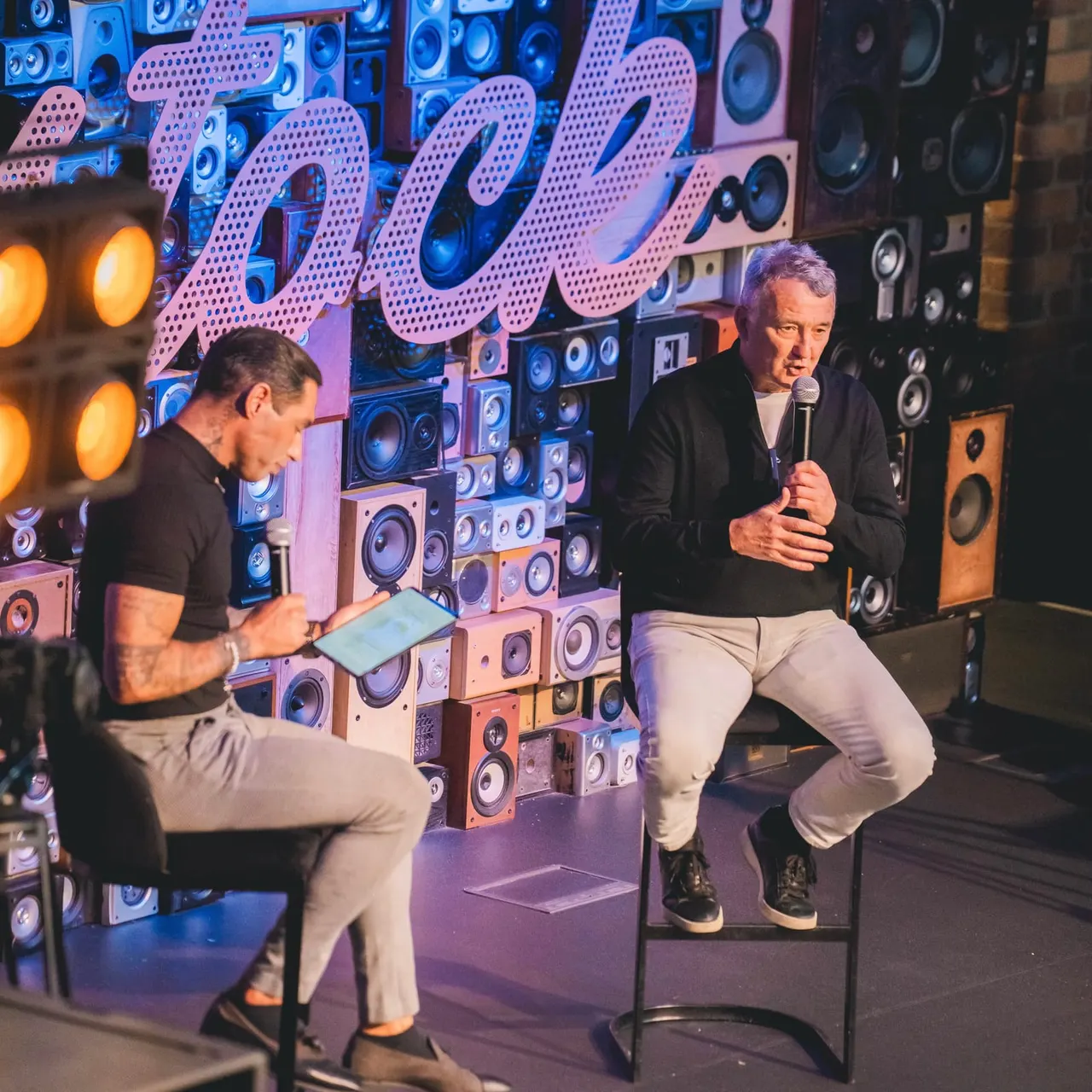 Two men perform on stage in front of a wall of illuminated speakers, one holding a microphone and the other holding a tablet device.