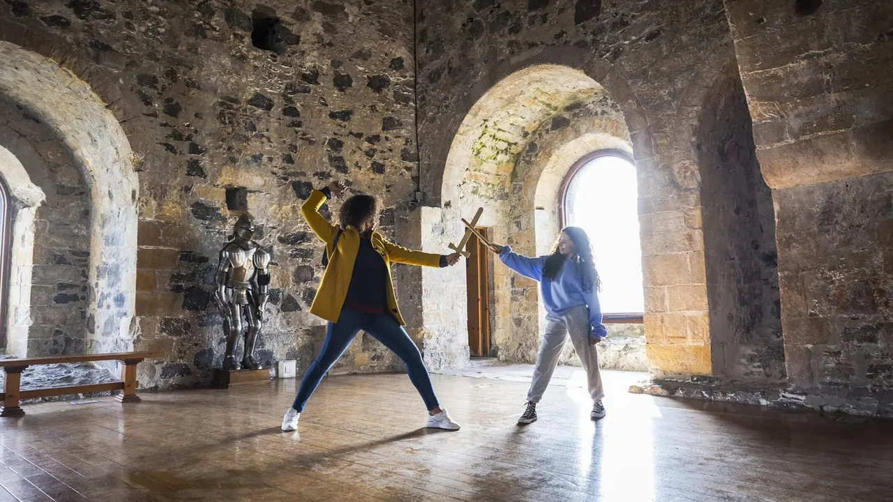 Two visitors practice sword fighting with wooden swords inside a historic stone fortress with arched ceilings and natural light.