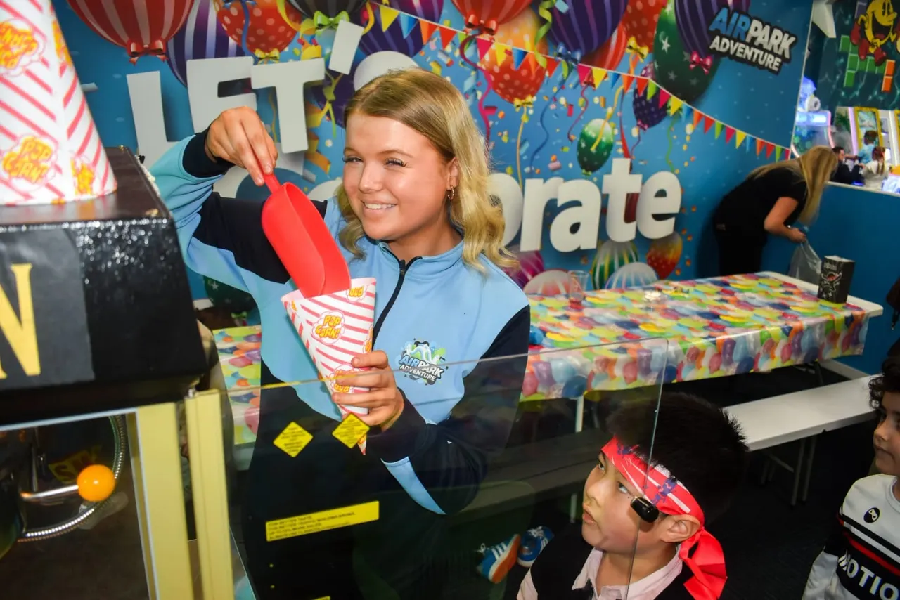 A girl in an Airpark Adventure uniform holds up a red novelty item while smiling at a younger child wearing a red pirate hat in a colourful party room.