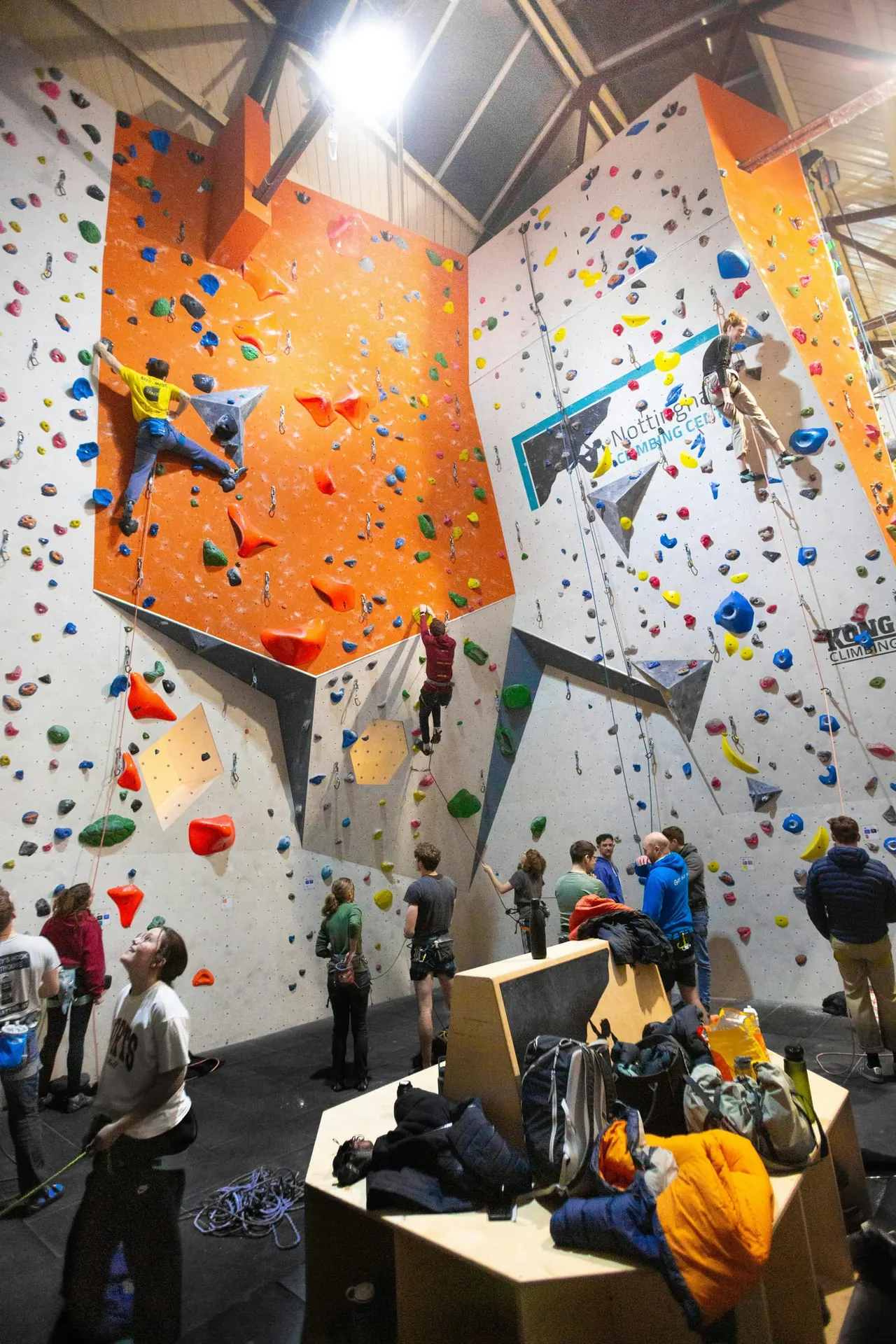 Children and adults rock climbing indoors on colourful holds across tall orange and white climbing walls.