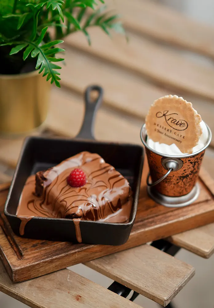Chocolate cake with raspberry on a cast-iron skillet next to a coffee cup, with potted herb on wooden board.