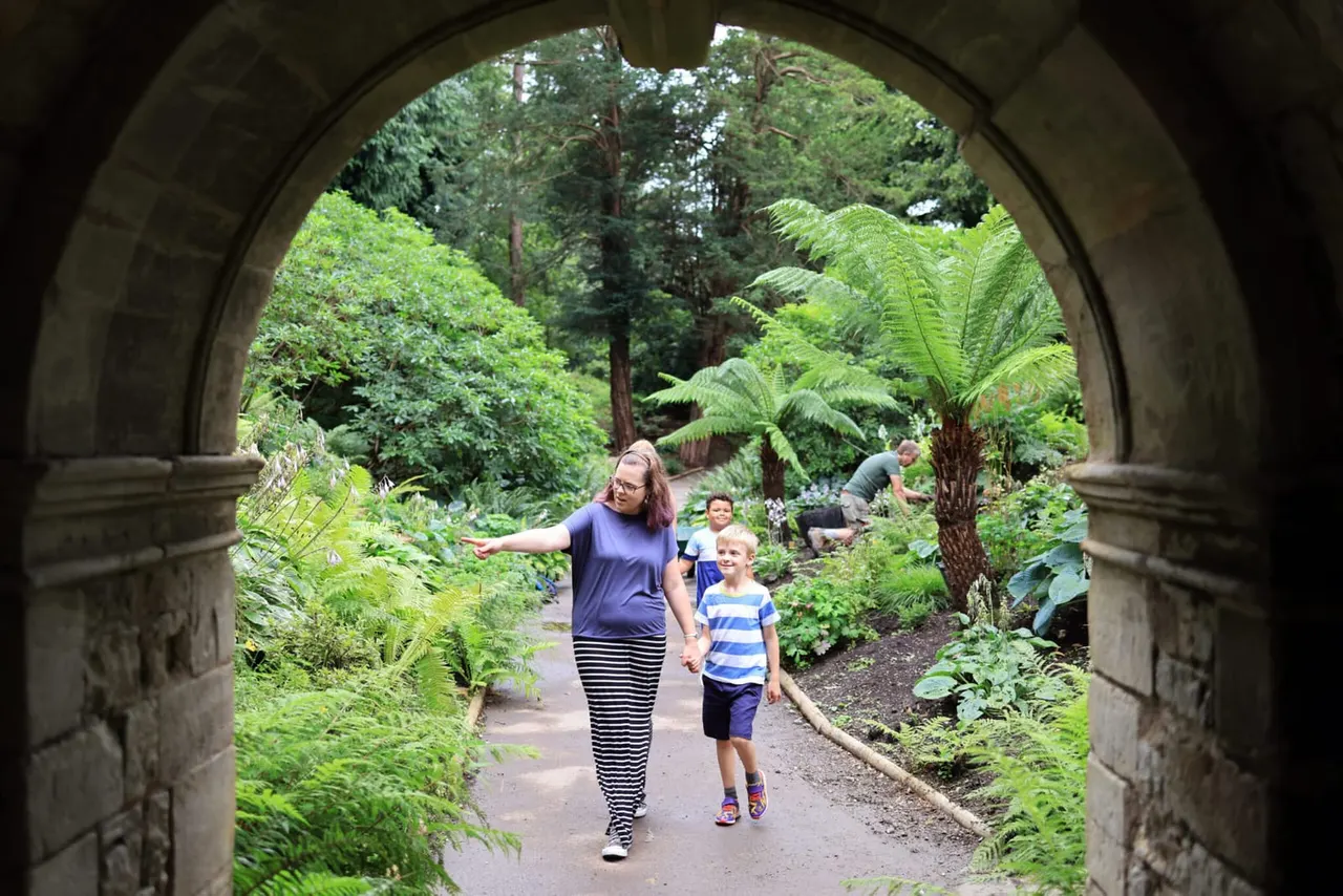 Woman and child walking hand-in-hand through a stone archway into a lush woodland garden with ferns and tall trees.
