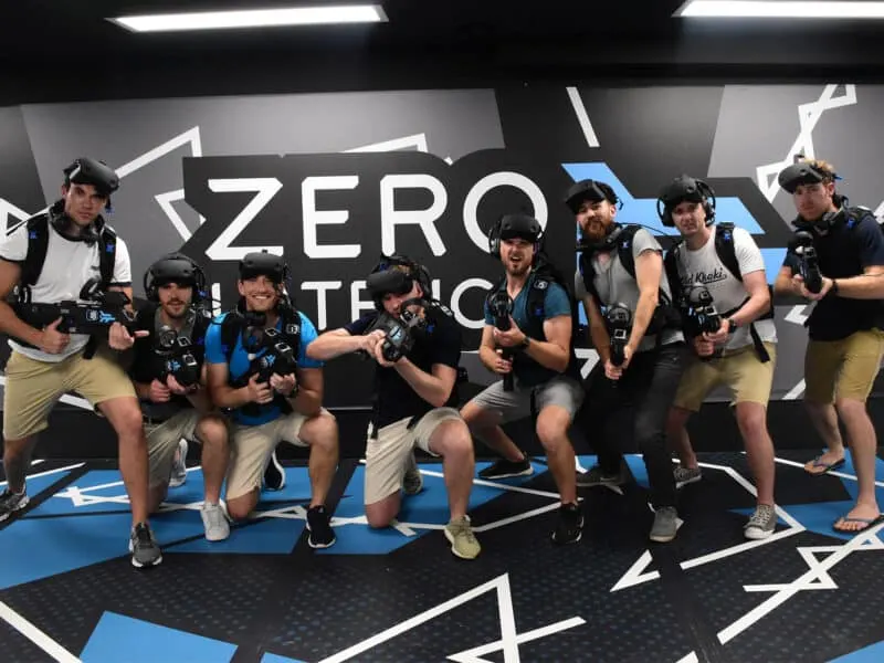 Group of people holding VR controllers in a gaming arena with "Zero Latency" branding visible on the wall.