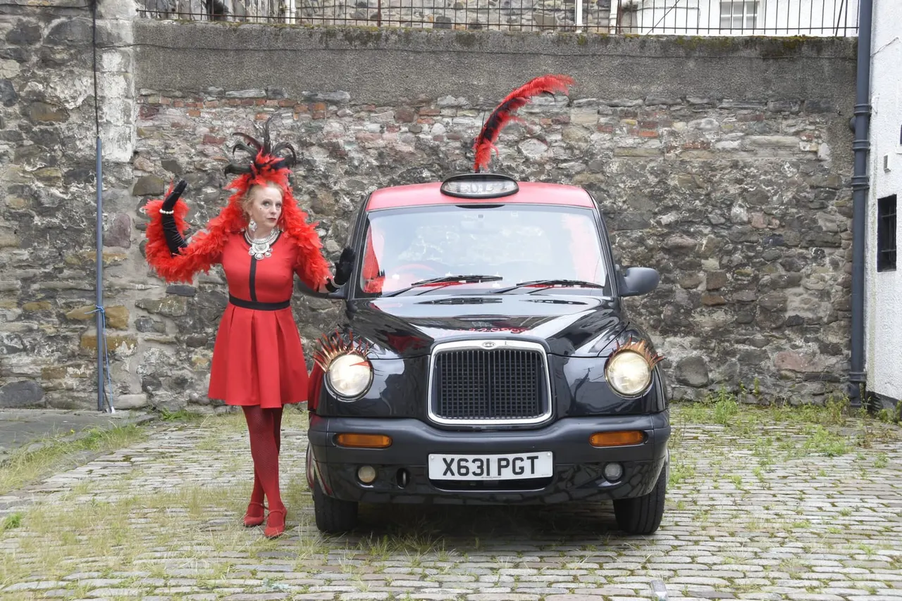 Woman in red outfit with feathered headpiece posing next to a black London taxi cab against a stone wall.