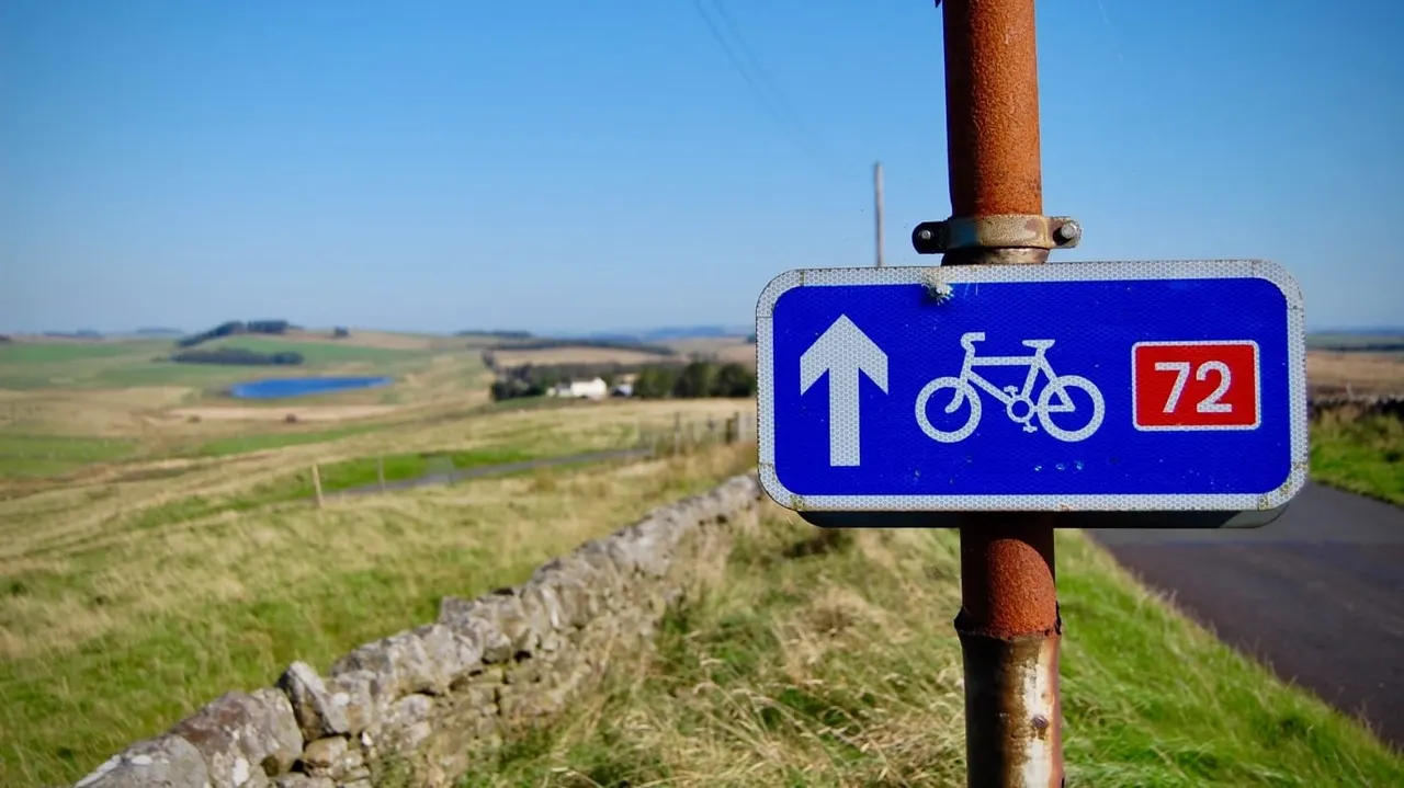 Cycling route marker sign on a rusted pole overlooking rolling countryside with fields, stone walls and distant trees.