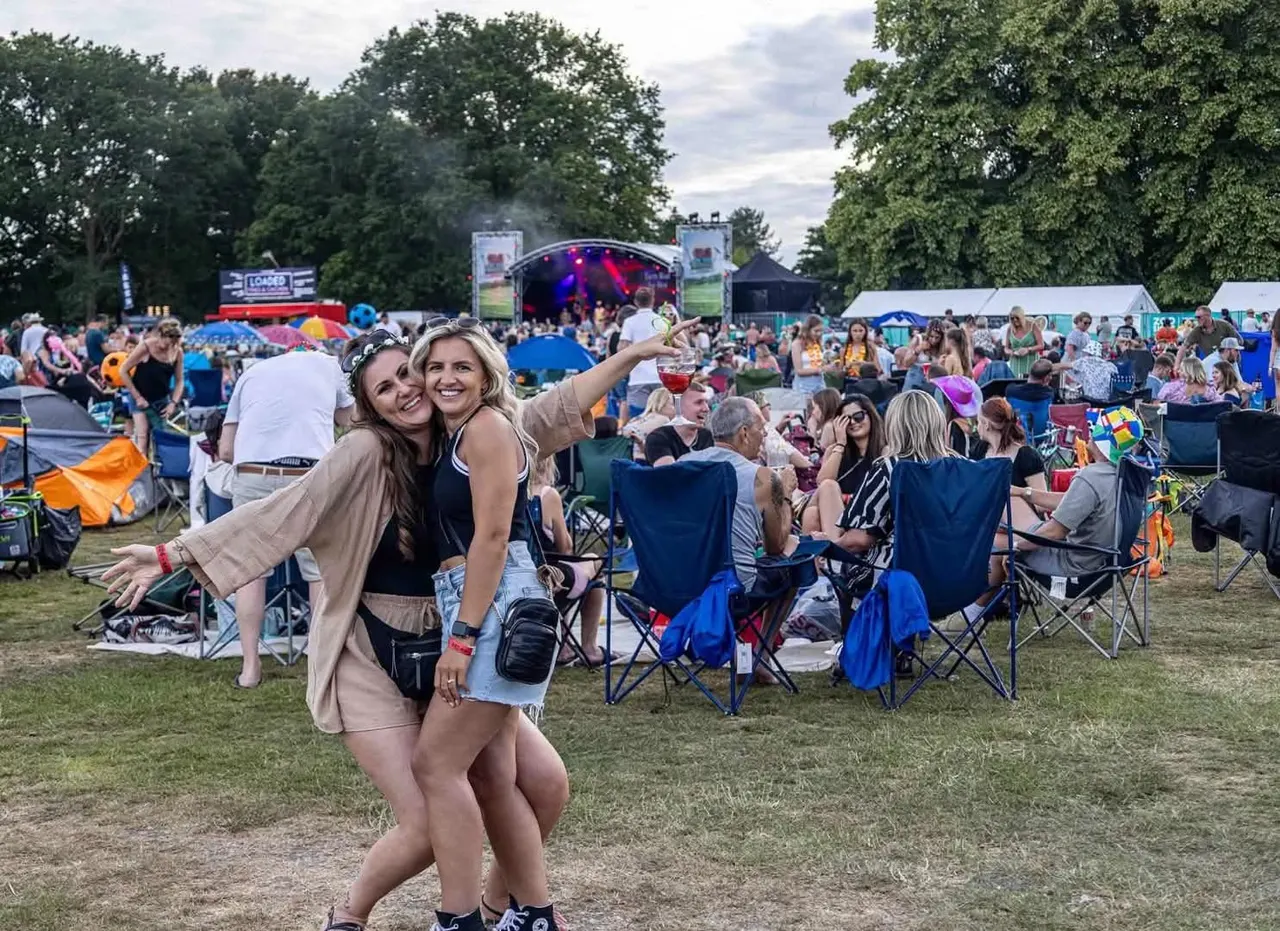 Two women pose for a selfie at an outdoor music festival with stage, crowds and camping chairs visible in the background.