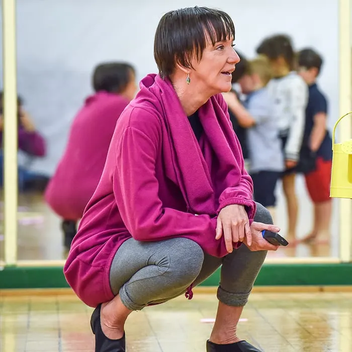 Woman in magenta jacket crouching indoors while children stand in background during a group activity.
