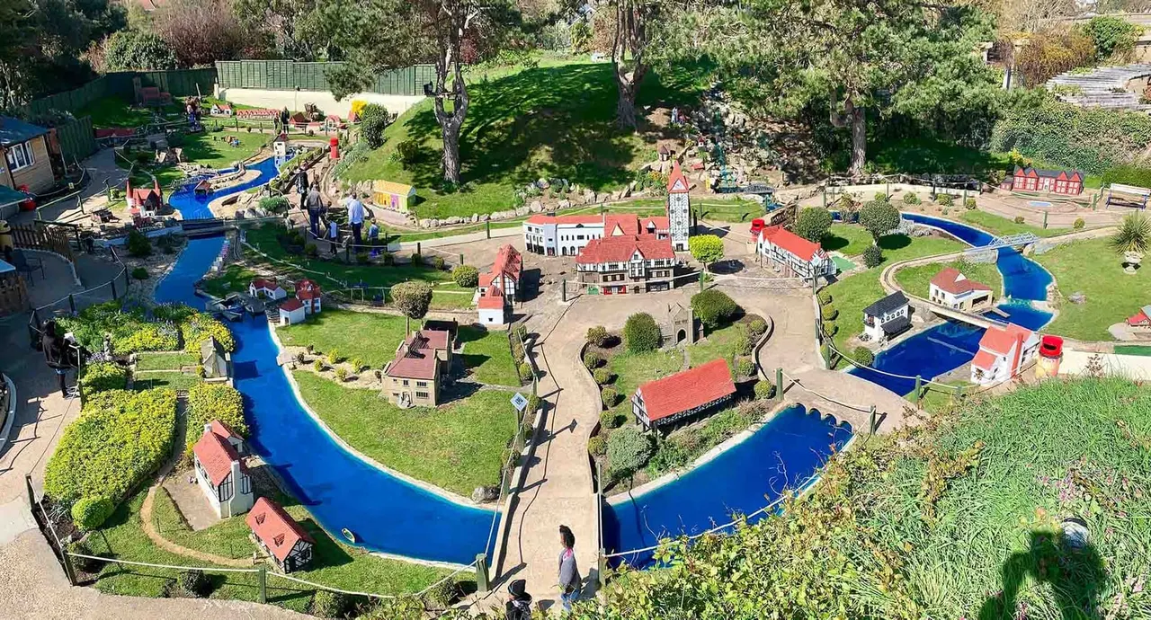 Aerial view of a miniature village attraction with blue waterways, red-roofed buildings, green lawns and visitors walking pathways.