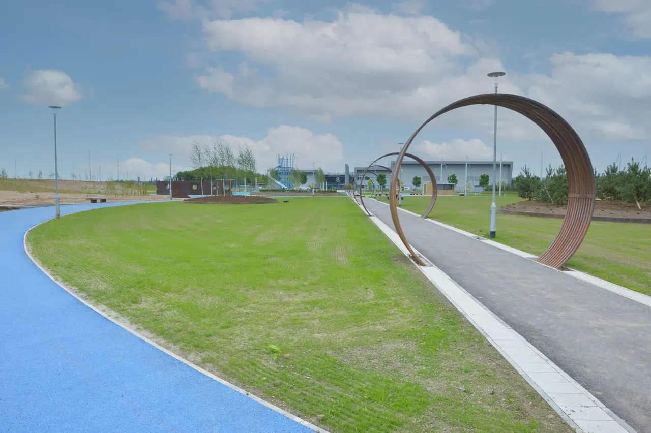 Modern playpark with large metal ring sculptures on a paved path, surrounded by green grass and play equipment in the distance.