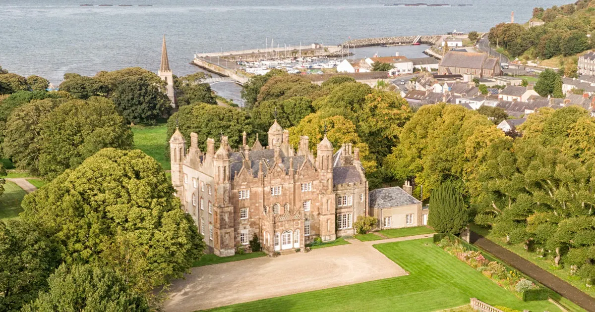 Aerial view of a stone castle surrounded by wooded grounds, with a marina and coastal town visible beyond.