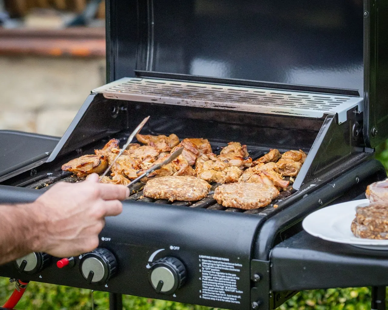 Person grilling burgers and chicken pieces on a barbecue outdoors.