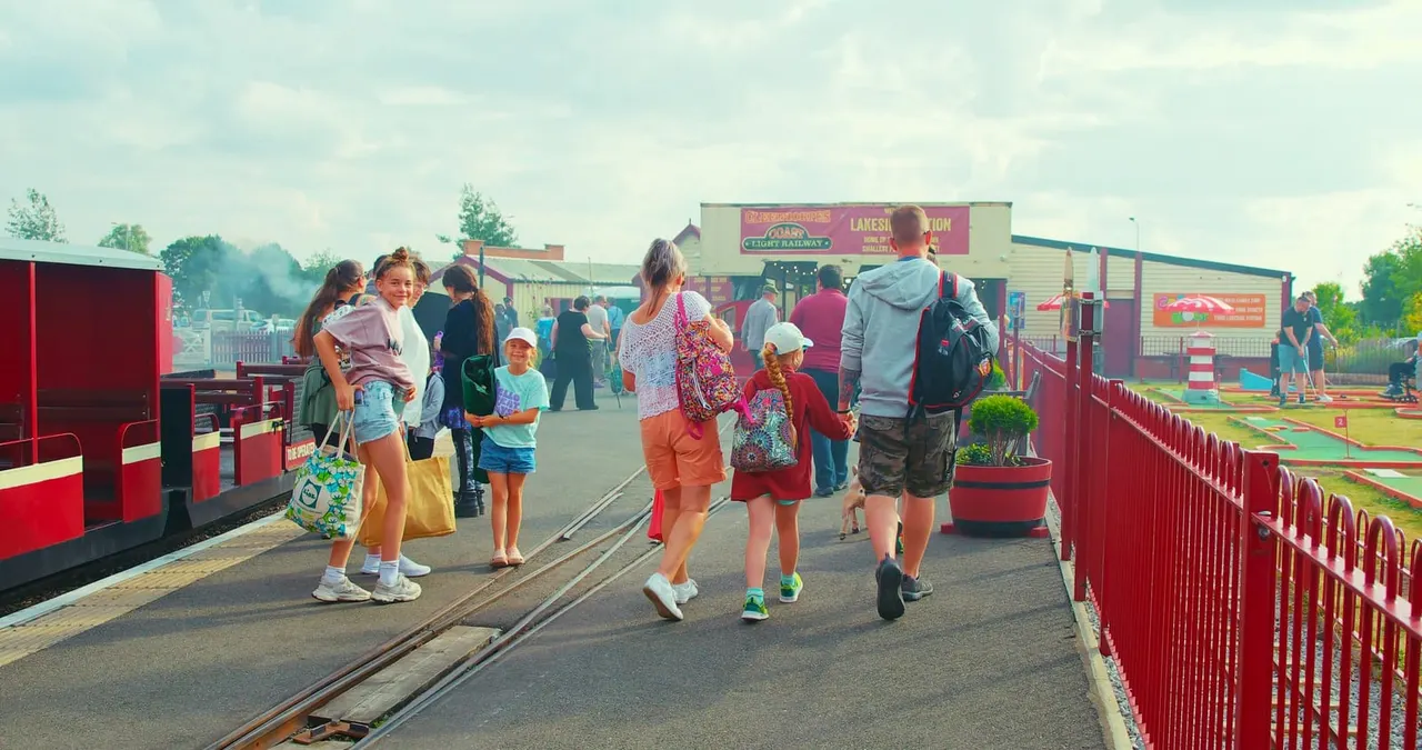Families and children walking along a railway platform next to red heritage train carriages and a miniature golf course.