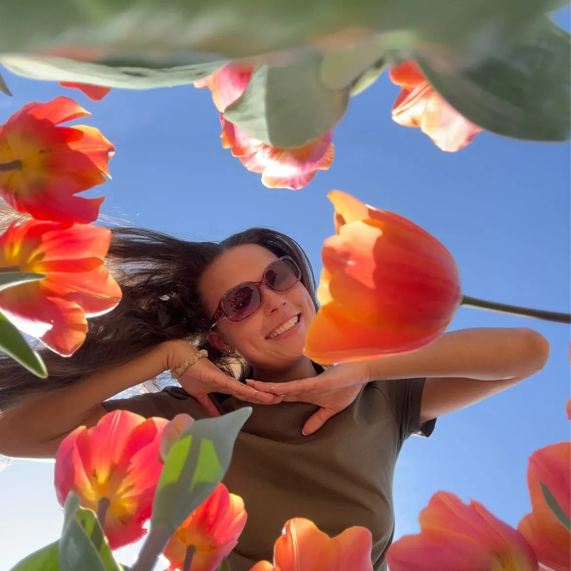Woman in sunglasses surrounded by bright red and orange tulips against a blue sky.