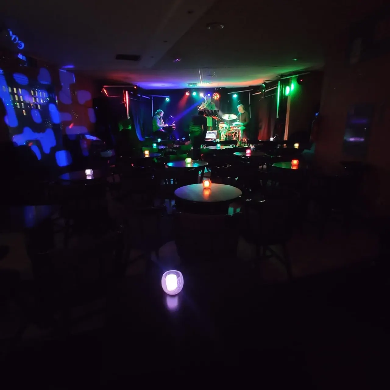 Live band performing on stage in a dark nightclub with colourful neon lighting and tables in the foreground.