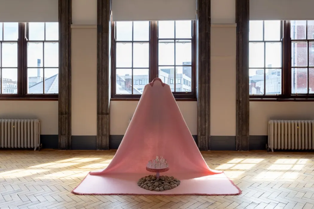 A large pink fabric cone sculpture inside a bright gallery space with tall windows overlooking a city skyline.