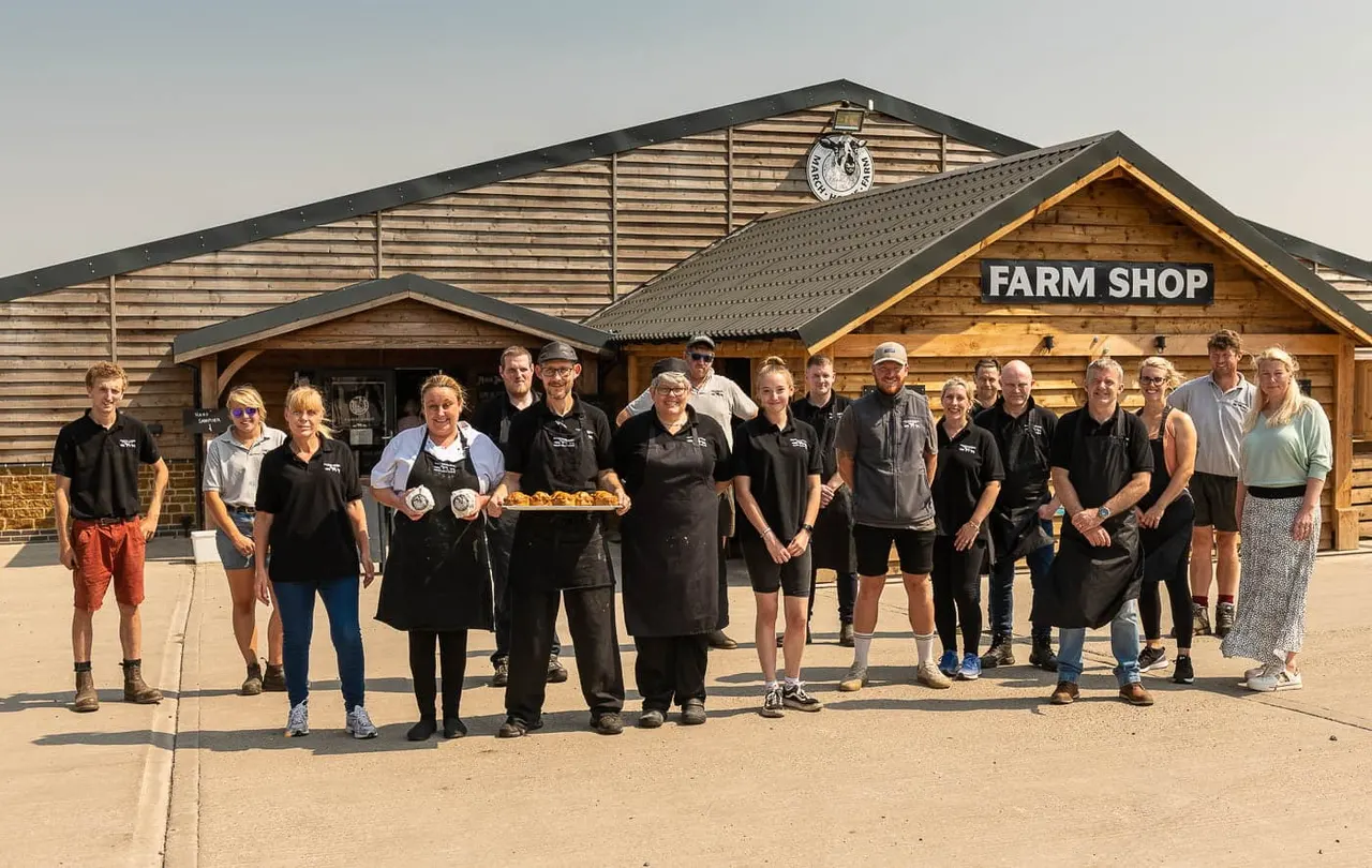 Staff and visitors stand together outside a wooden farm shop building on a sunny day.