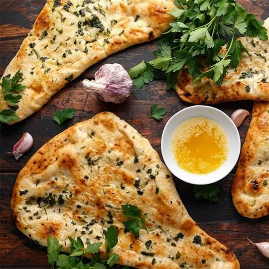 Herb-topped flatbreads with garlic cloves, fresh parsley and a bowl of olive oil on a wooden table.