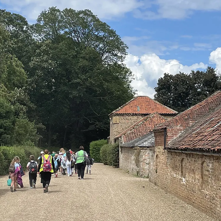 Visitors walking along a gravel path towards a historic stone and brick building with terracotta roof tiles, surrounded by trees.