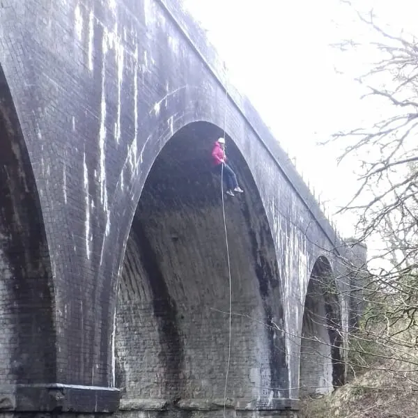 Person abseiling down a large stone railway viaduct arch with bare trees visible alongside.