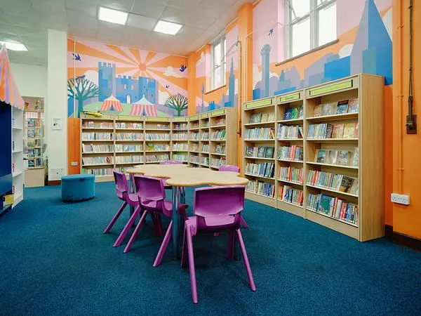 Colourful children's library space with purple chairs at a reading table, bookshelves, and vibrant murals.