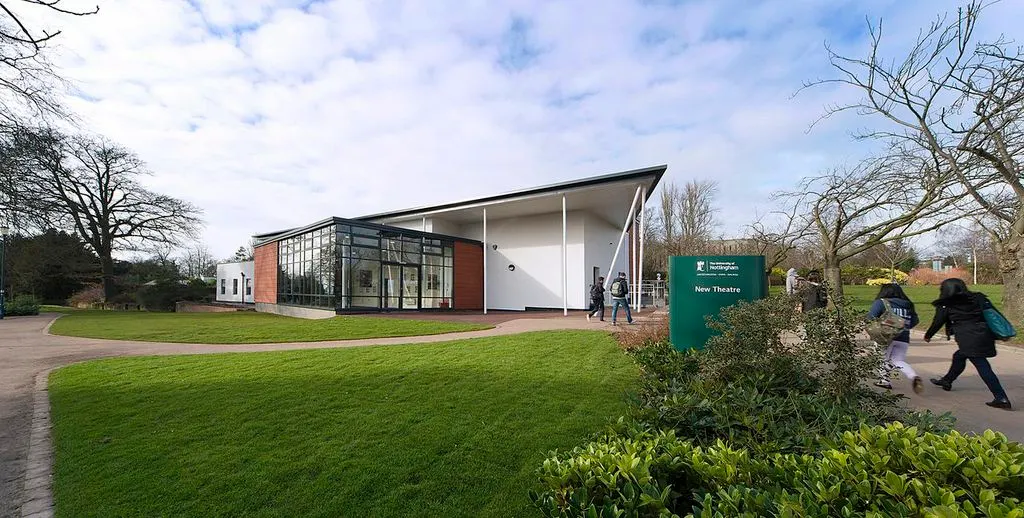 Modern brick and white theatre building with large windows set in landscaped grounds, visitors walking towards entrance on paved path.