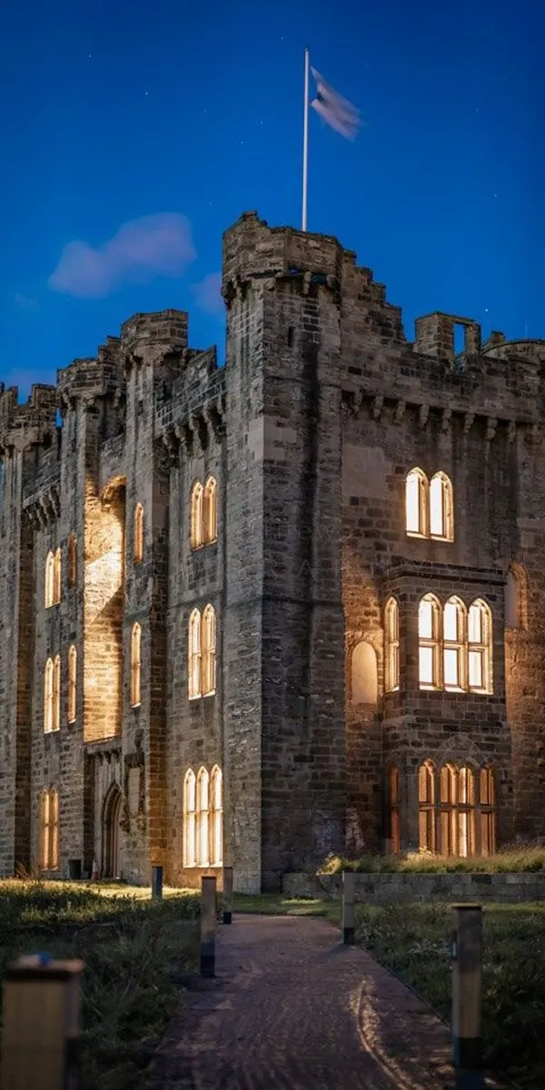 Historic stone castle with illuminated windows at dusk, flag flying from central tower, against deep blue twilight sky.