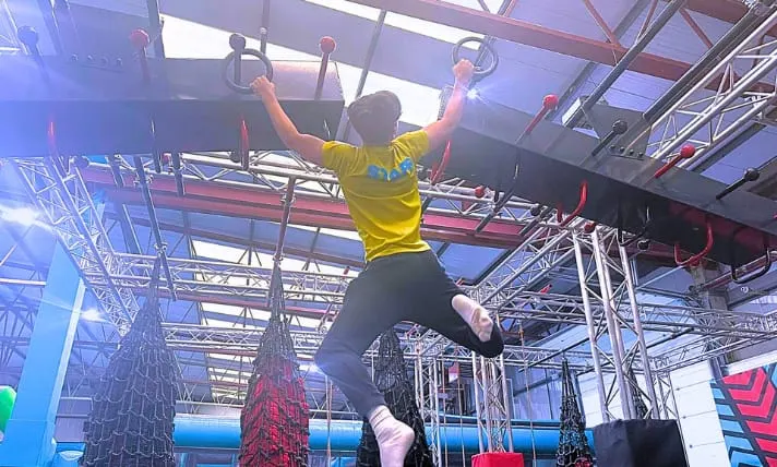 Person in yellow shirt hanging from overhead bars on an indoor obstacle course with metal framework and netting.