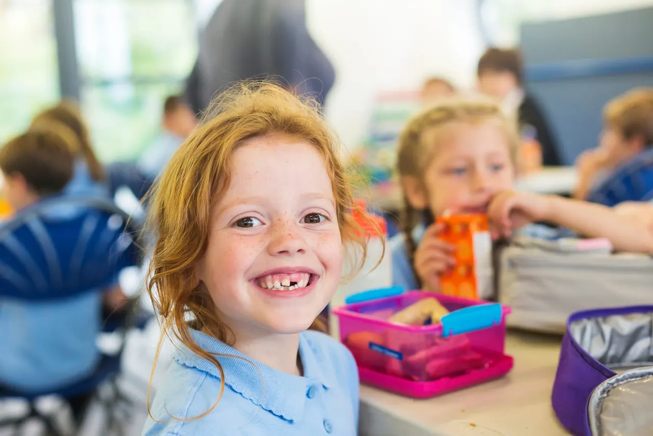 Young child with red hair smiling at camera during a classroom arts and crafts activity with other children.
