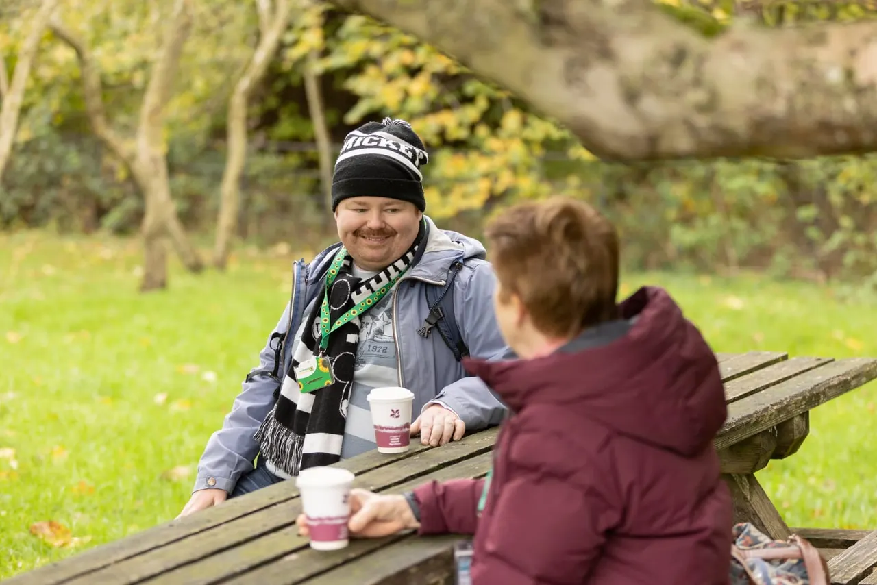 Two people sitting at a wooden picnic table in a wooded garden, holding hot drinks and smiling.