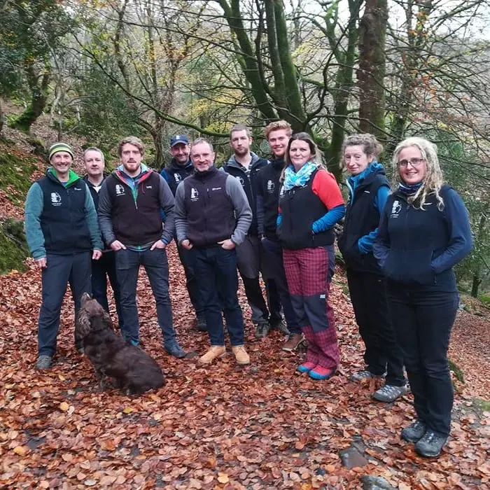 A group of eleven people wearing outdoor activity clothing stand together on a woodland path covered with fallen leaves, bare trees visible behind them.