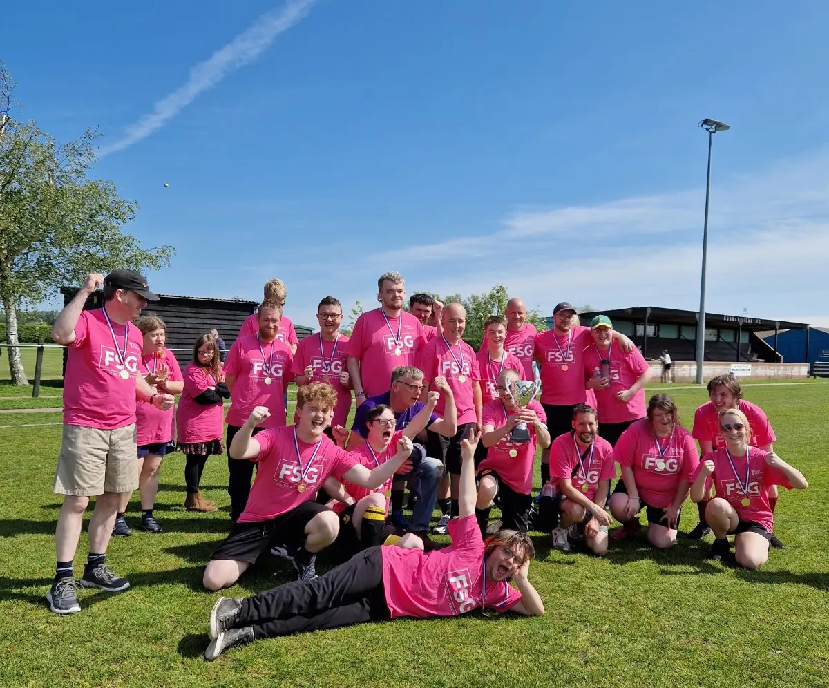 A large group of people wearing matching pink charity t-shirts pose together on a sunny grass field at a sports facility.