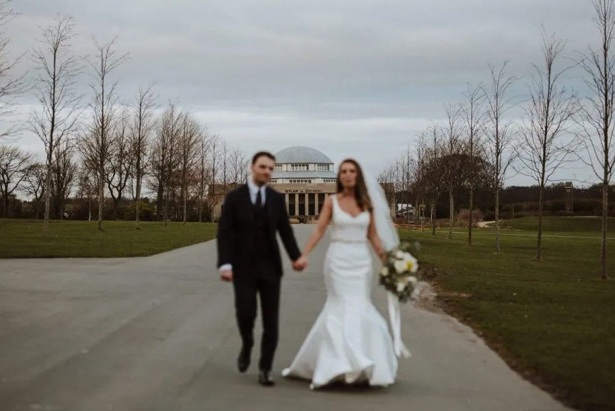 Bride and groom walking hand in hand along a paved path lined with bare trees, with a historic building in the background.