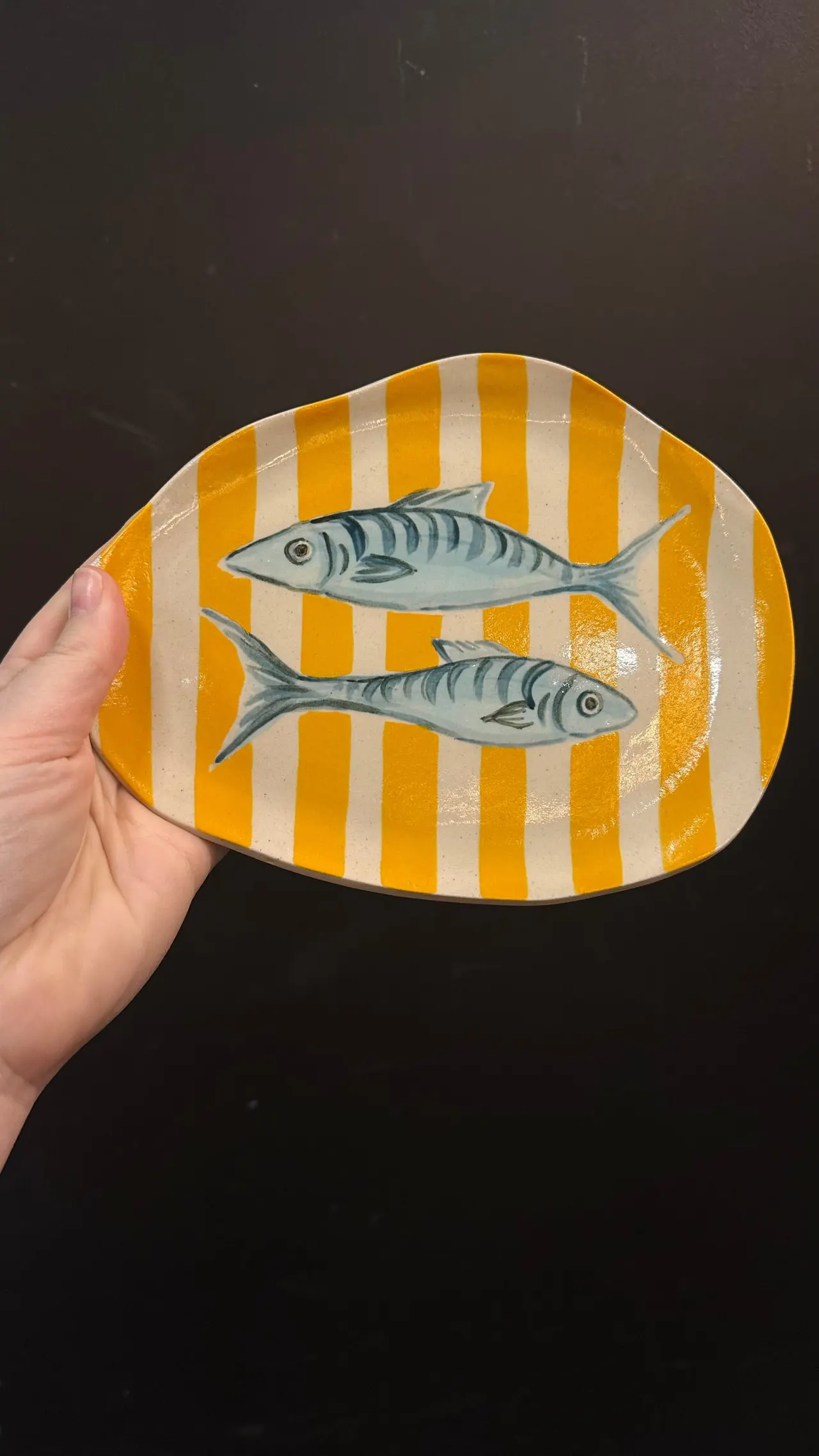 Hand holding a ceramic plate with yellow and white stripes decorated with two illustrated fish.