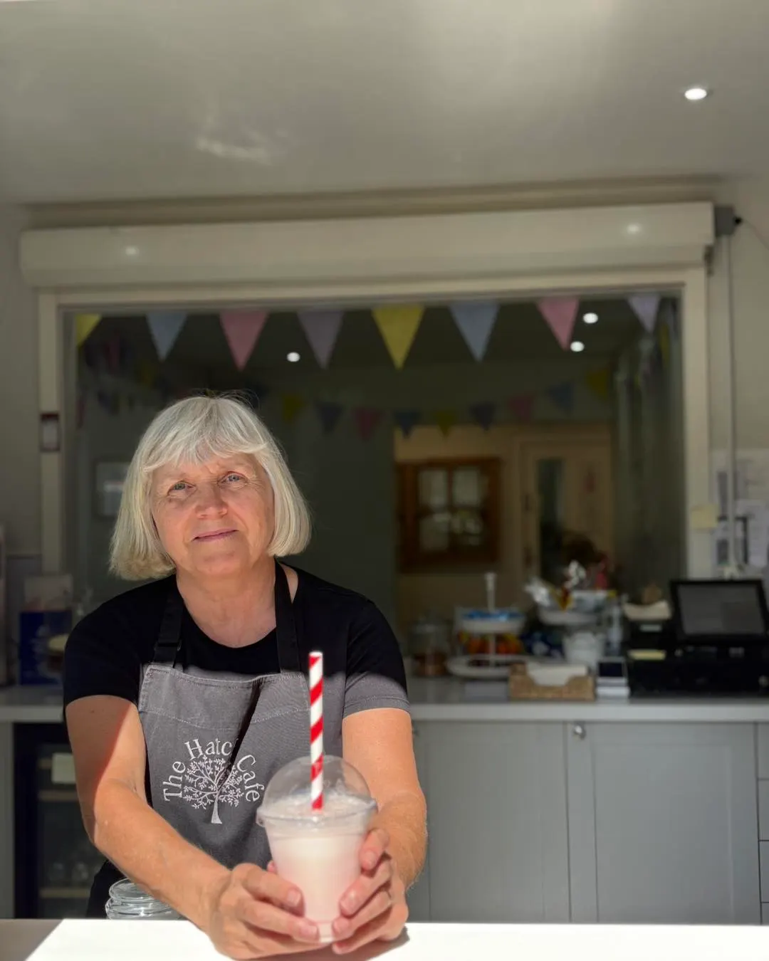 Woman in apron holding a milkshake behind a cafe counter with bunting and kitchen visible.