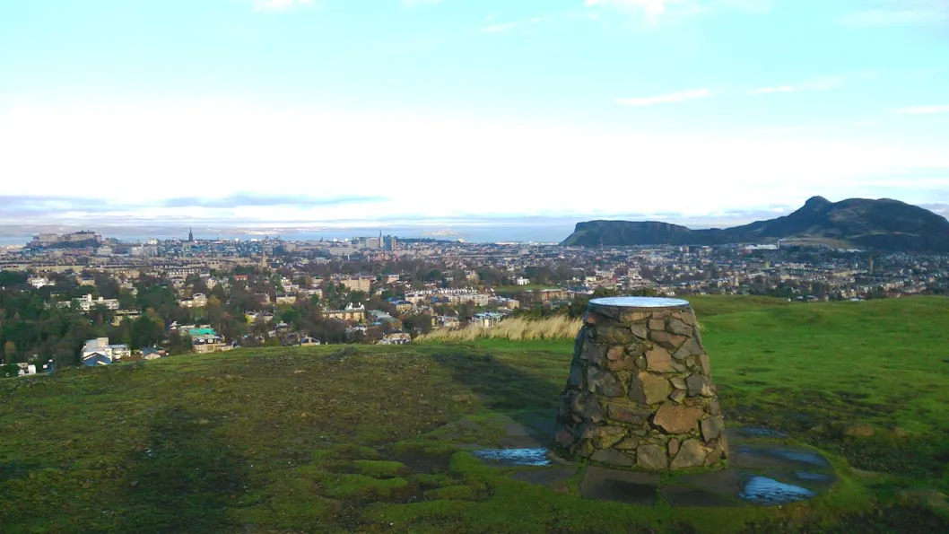 Stone cairn on grassy hillside overlooking Edinburgh cityscape with distant mountains and blue sky.