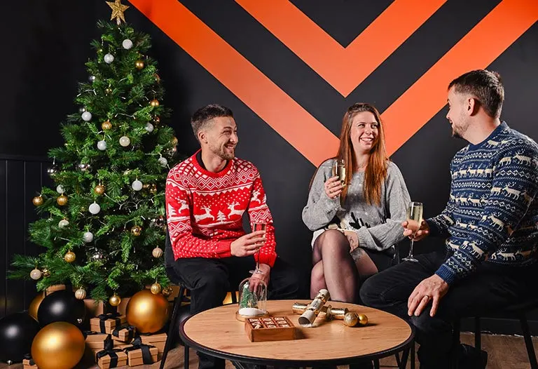 Three people in festive jumpers socialising by a decorated Christmas tree with drinks and games.