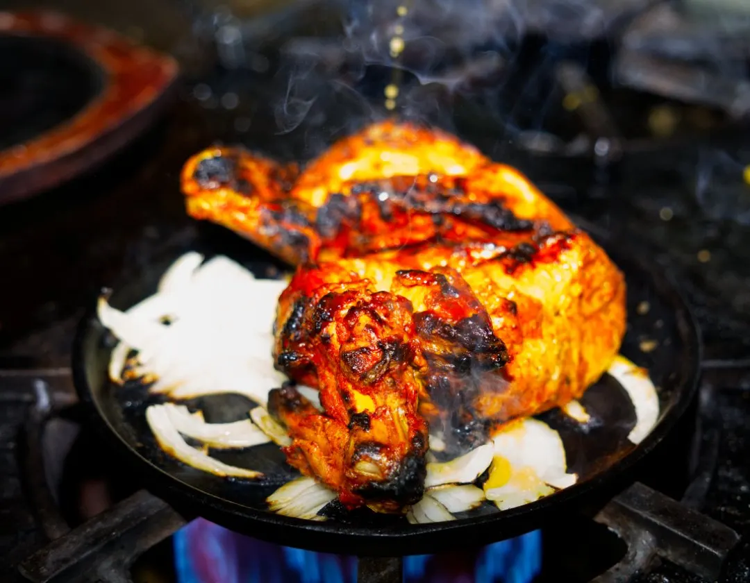 Sizzling tandoori chicken on a cast-iron plate with sliced onions and steam rising, served over an open flame.