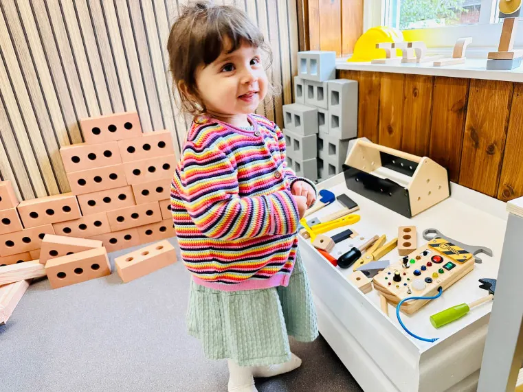 Toddler in colourful striped jumper playing with wooden building blocks and educational toys at an indoor play venue.