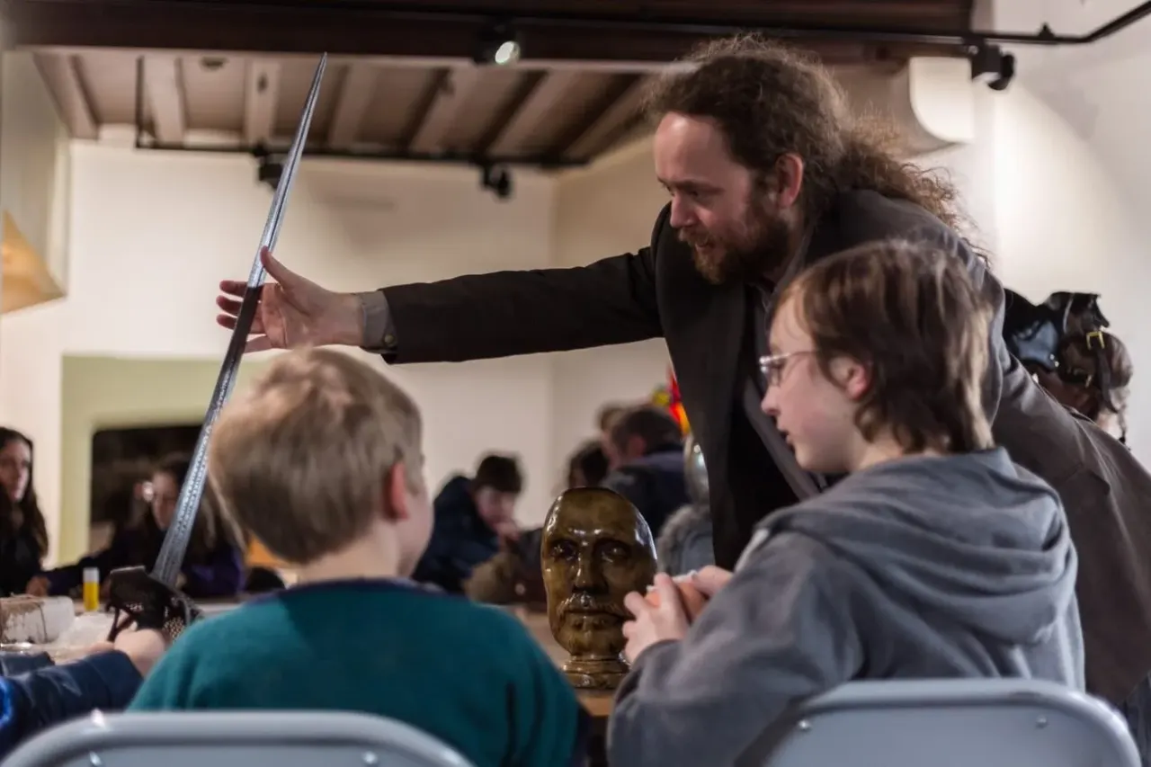 A bearded instructor demonstrates a sword and bronze head sculpture to children seated at a table during an indoor activity session.