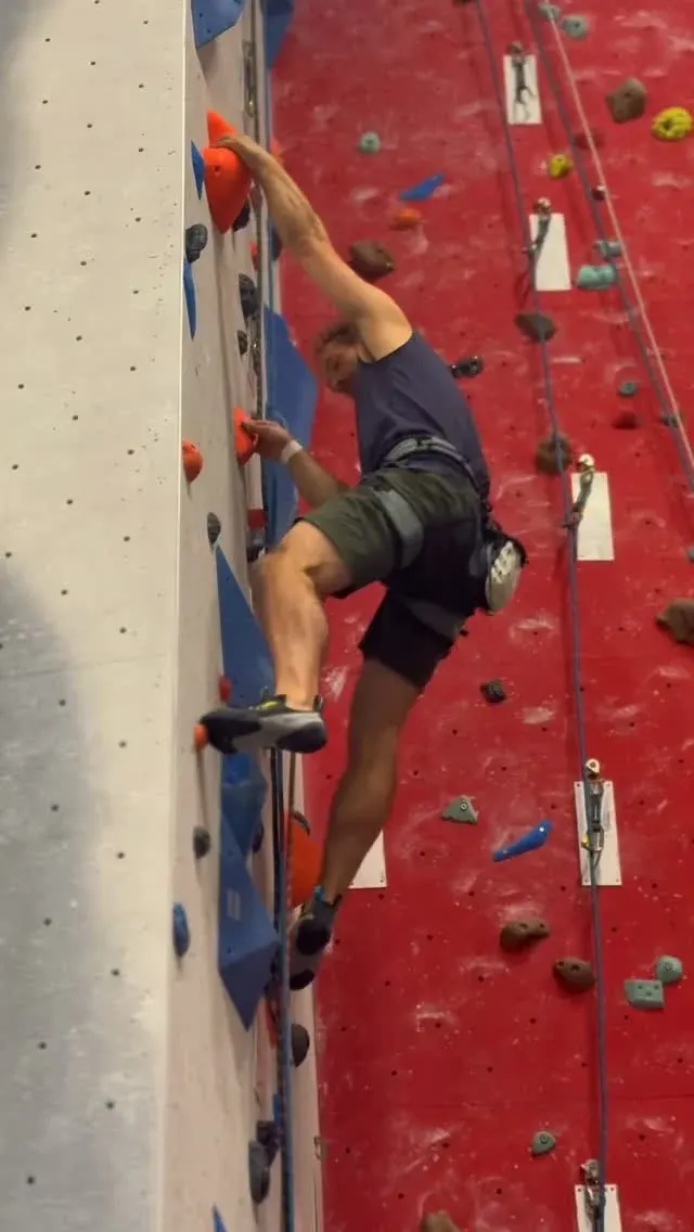 Person indoor rock climbing on a red wall with coloured holds and safety rope.