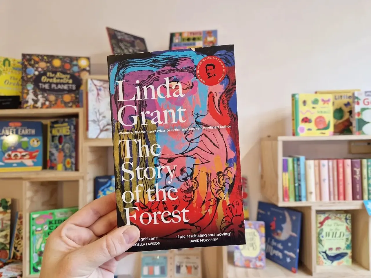 A hand holding Linda Grant's "The Story of the Forest" book in front of shelving units filled with colourful books.