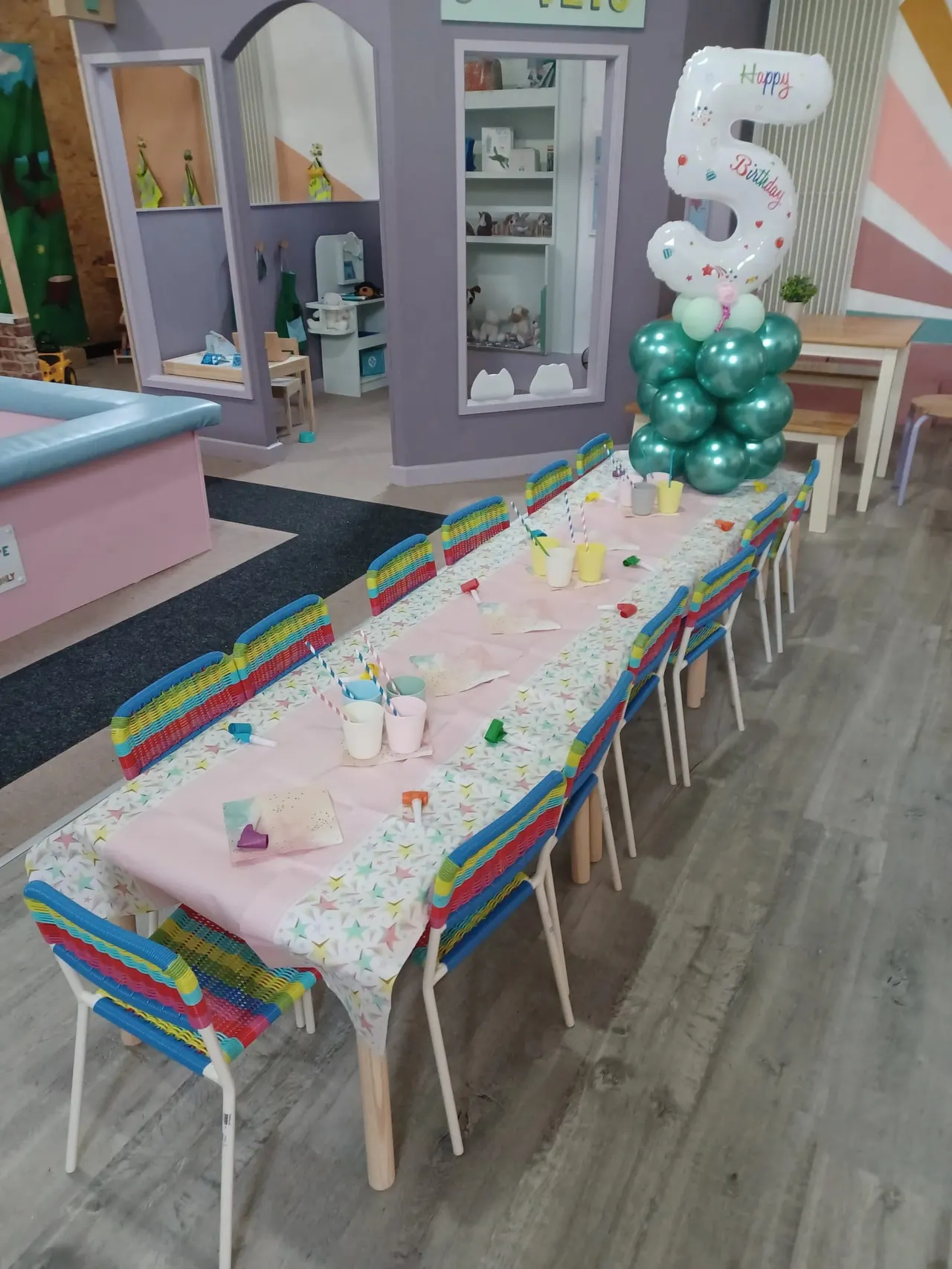 Children's party room with decorated table set with cups and craft supplies, green balloon display, and birthday decorations.