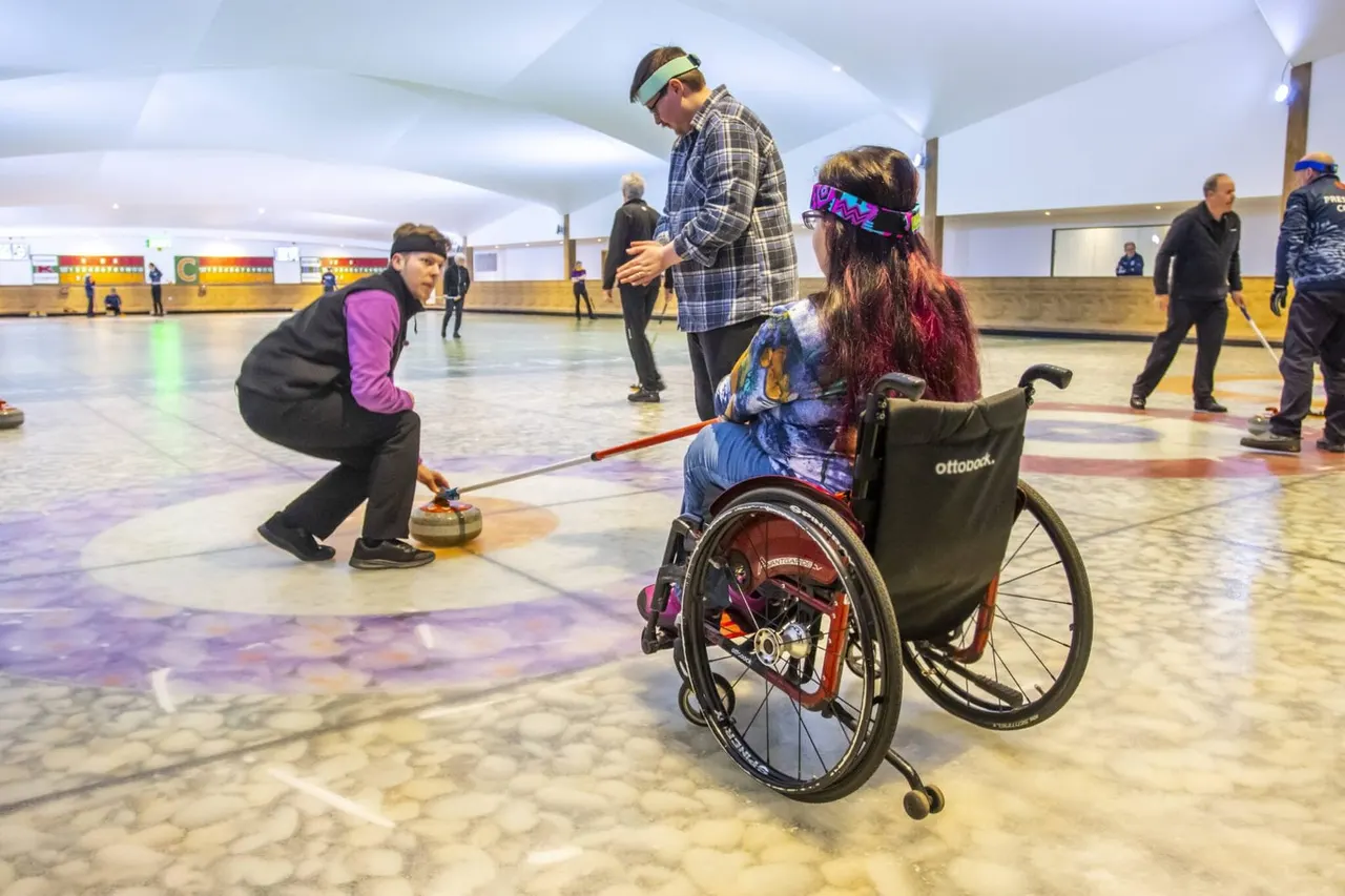 Wheelchair curling player delivers stone whilst teammates and instructor watch on indoor ice rink.