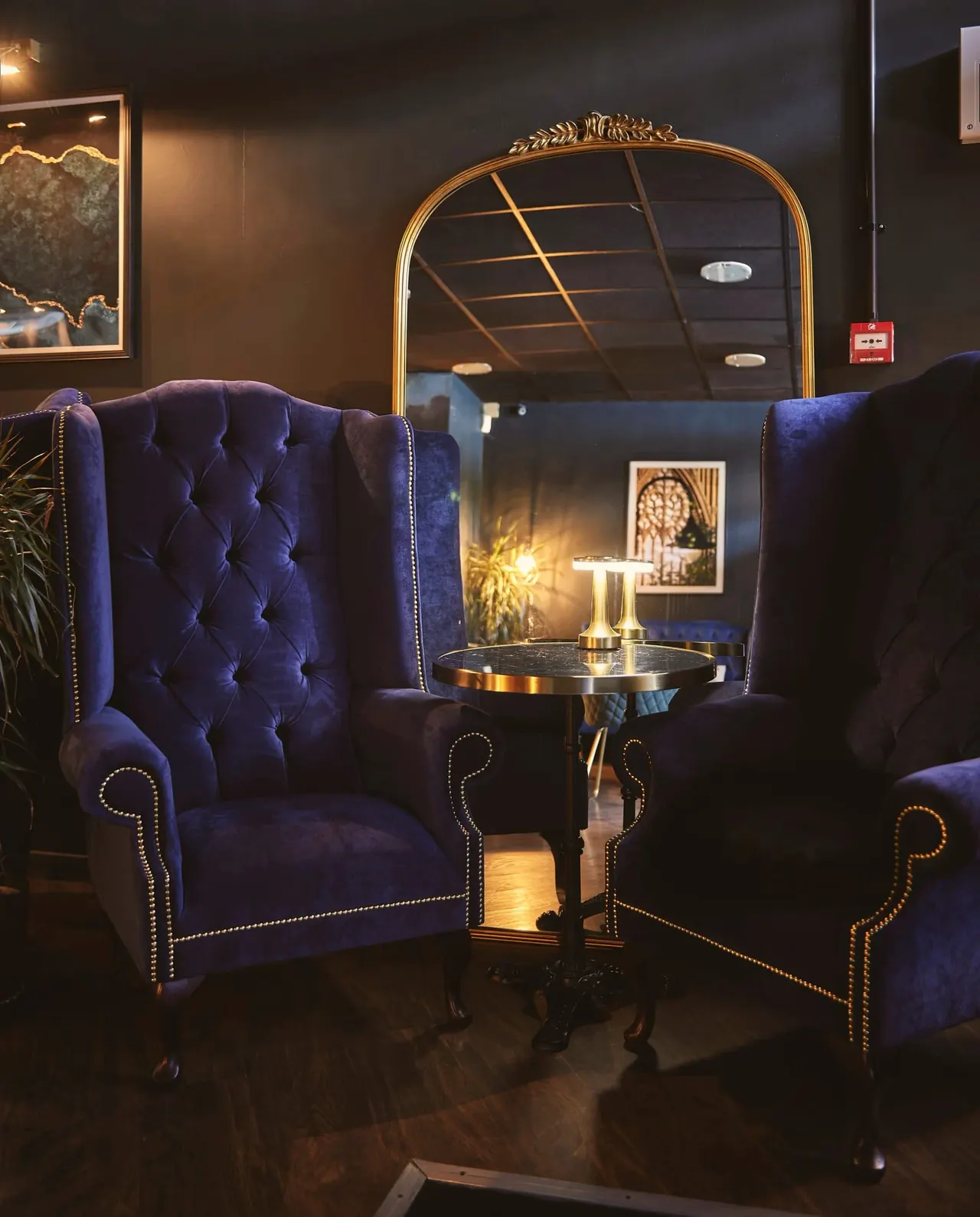 Luxurious lounge seating area with deep blue velvet wingback chairs and a small round table, lit by warm golden lighting and a large arched mirror.
